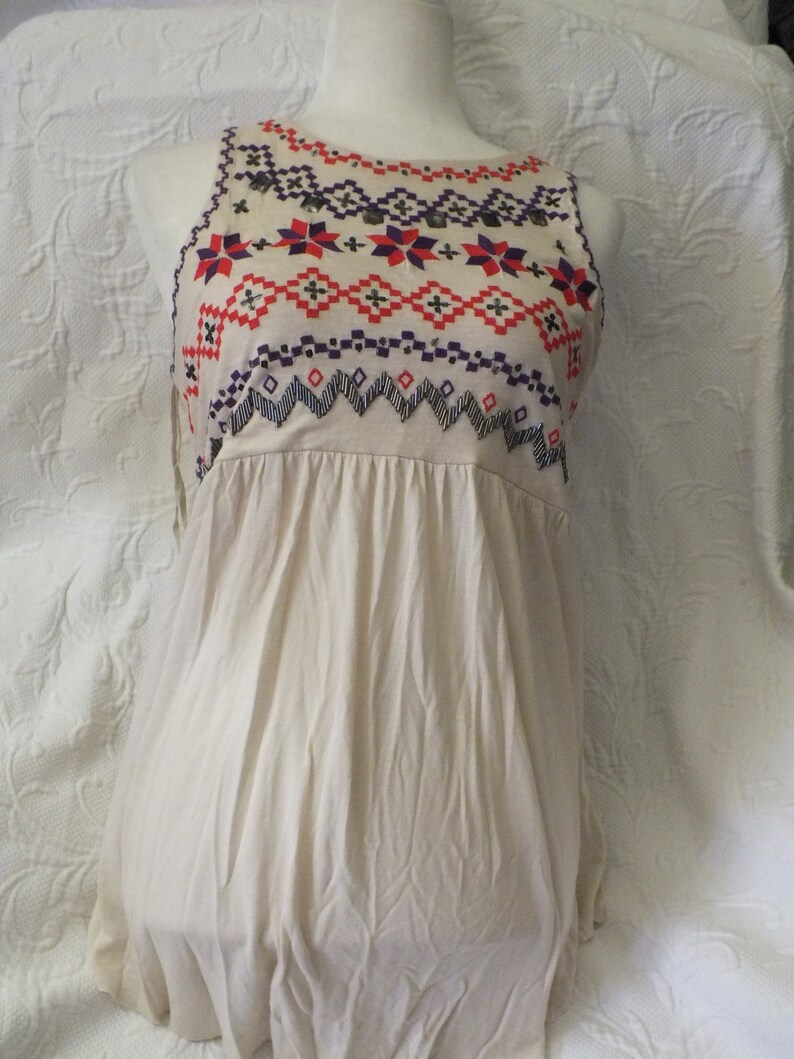May include: A sleeveless, cream-colored, cotton dress with a geometric pattern embroidered on the neckline in shades of red, orange, purple, and black.