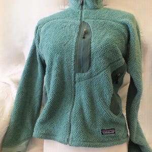 May include: A teal fleece jacket with a full-length zipper and a high collar. The jacket features a chest pocket with a zipper and two side pockets. The Patagonia logo is visible on the lower left side. The jacket is displayed on a white mannequin.
