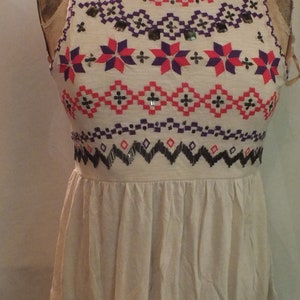 May include: A white sleeveless top with a geometric pattern embroidered in pink, purple, and black. The pattern includes stars and diamonds. The top has a gathered skirt.