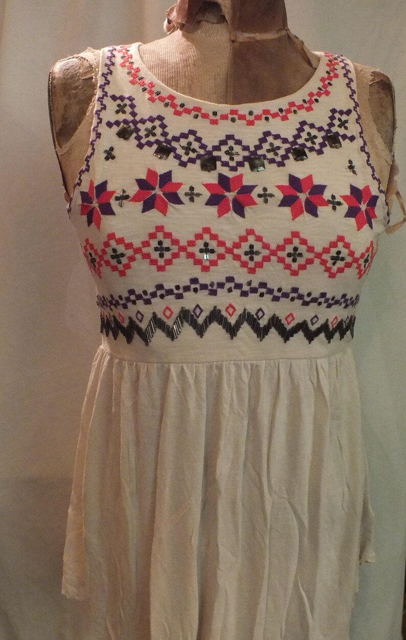 May include: A white sleeveless top with a geometric pattern embroidered in pink, purple, and black. The pattern includes stars and diamonds. The top has a gathered skirt.