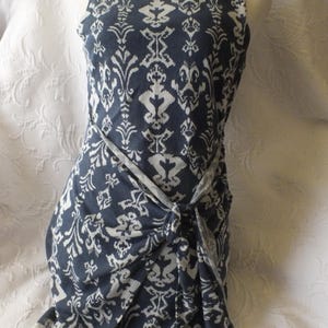 May include: Sleeveless navy blue dress with a white damask pattern. The dress features a wrap-style design with a knotted waist detail. The fabric appears soft and drapes elegantly, creating a flattering silhouette.