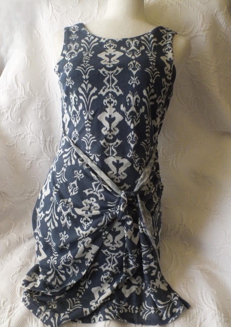 May include: Sleeveless navy blue dress with a white damask pattern. The dress features a wrap-style design with a knotted waist detail. The fabric appears soft and drapes elegantly, creating a flattering silhouette.