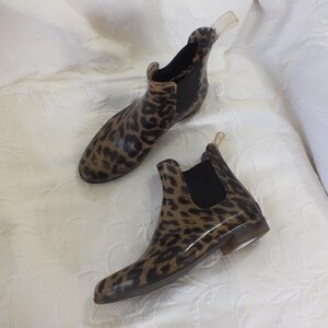 May include: A pair of ankle-high rain boots with a leopard print pattern. The boots have a brown and black design, with black elastic side panels and a clear pull tab. The boots are made of a glossy material.