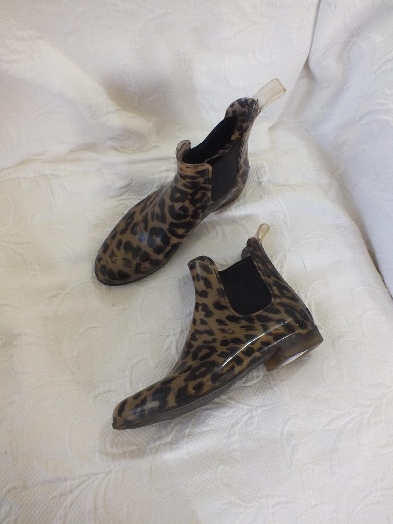 May include: A pair of ankle-high rain boots with a leopard print pattern. The boots have a brown and black design, with black elastic side panels and a clear pull tab. The boots are made of a glossy material.
