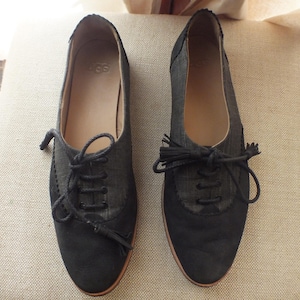 May include: A pair of dark charcoal and black lace-up shoes featuring a textured fabric upper, smooth toe caps, and black laces with tassel ends. The interior insoles are tan and feature the UGG brand logo embossed in the center of the heel area.