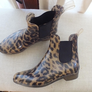 May include: A pair of ankle-high rain boots with a leopard print pattern. The boots have a brown and black design, a black elastic panel on the sides, and a clear pull tab at the back. The boots are made of a glossy material.