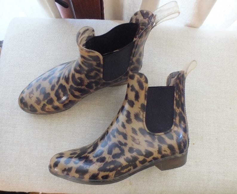 May include: A pair of ankle-high rain boots with a leopard print pattern. The boots have a brown and black design, a black elastic panel on the sides, and a clear pull tab at the back. The boots are made of a glossy material.
