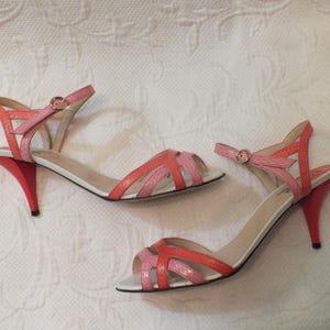 May include: A pair of open-toe sandals with a red, pink, and white color scheme. The sandals have a strap around the ankle and a small heel. The heels are approximately 3 inches tall.