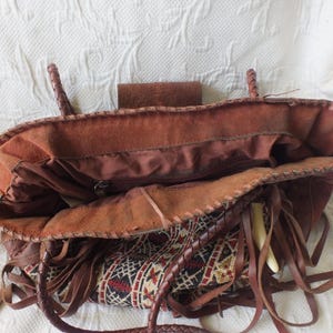 May include: Open view of a brown leather tote bag with braided handles and fringe detailing. The bag features a patterned fabric insert with red, black, and cream-colored geometric designs. The interior is visible, showing a zippered pocket.