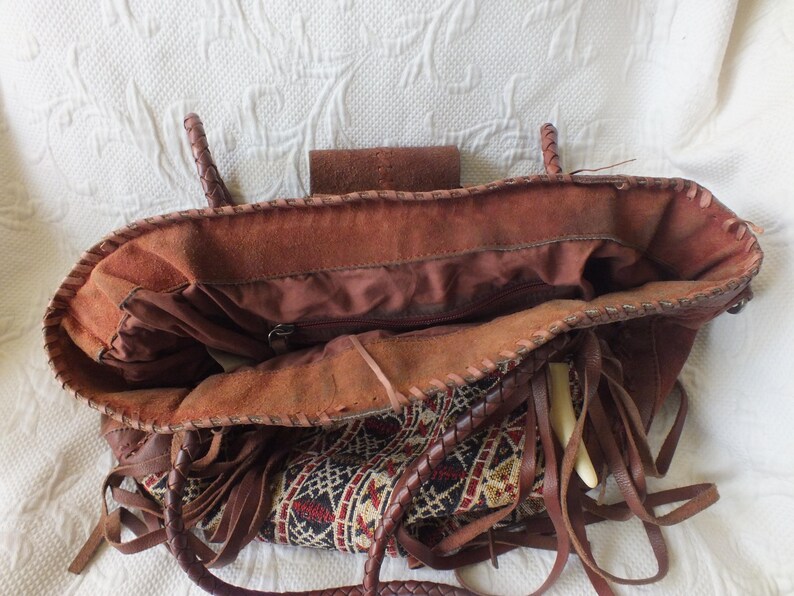 May include: Open view of a brown leather tote bag with braided handles and fringe detailing. The bag features a patterned fabric insert with red, black, and cream-colored geometric designs. The interior is visible, showing a zippered pocket.