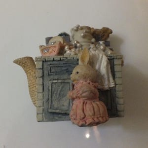 May include: A ceramic teapot shaped like a kitchen with a bunny peeking out of a cabinet door. The teapot is decorated with a white and gray kitchen cabinet, a pink bunny in a pink dress, and a white towel hanging from the cabinet door.