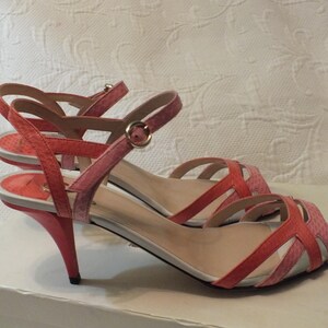 May include: A pair of coral and pink heeled sandals. The open-toe shoes have thin straps and a buckle closure. The heels are a curved, stiletto style. The sandals are displayed on a white surface.