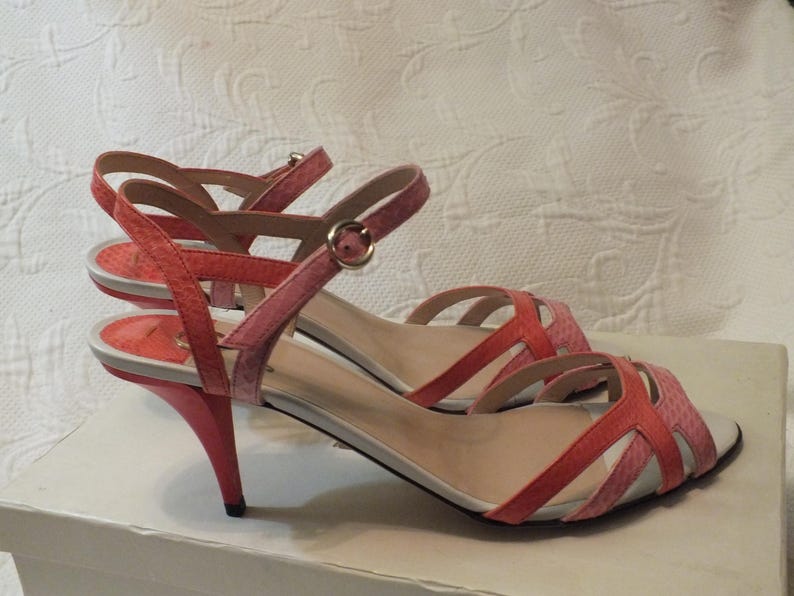 May include: A pair of coral and pink heeled sandals. The open-toe shoes have thin straps and a buckle closure. The heels are a curved, stiletto style. The sandals are displayed on a white surface.