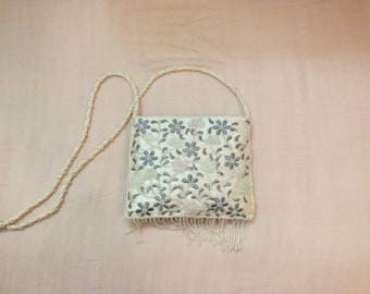 Puede incluir: Un bolso de noche blanco con cuentas, con un diseño floral y un ribete de flecos. El bolso tiene una correa larga.