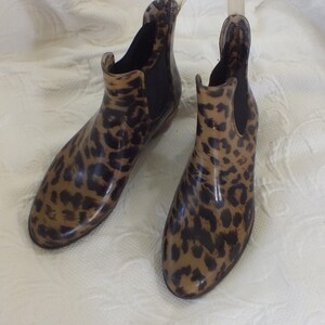May include: A pair of ankle-high rain boots with a leopard print pattern. The boots are a mix of tan and brown, with black accents. They have a glossy finish and a pull tab at the back. The boots are designed for wet weather.