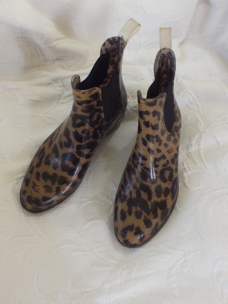 May include: A pair of ankle-high rain boots with a leopard print pattern. The boots are a mix of tan and brown, with black accents. They have a glossy finish and a pull tab at the back. The boots are designed for wet weather.