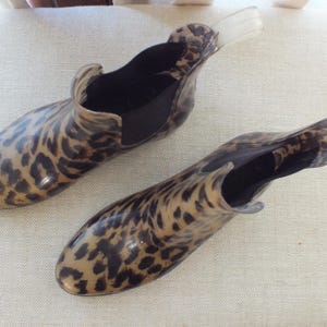 May include: A pair of clear, ankle-high rain boots with a leopard print pattern. The boots have a black elastic panel on the sides and a clear pull tab at the back. The boots are made of a shiny, waterproof material.