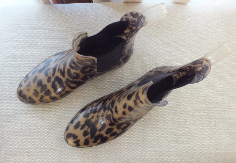 May include: A pair of clear, ankle-high rain boots with a leopard print pattern. The boots have a black elastic panel on the sides and a clear pull tab at the back. The boots are made of a shiny, waterproof material.