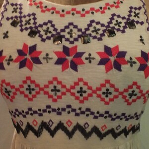 May include: A white sleeveless dress with a geometric pattern embroidered in red, purple, and black thread. The dress has a gathered skirt and a round neckline.