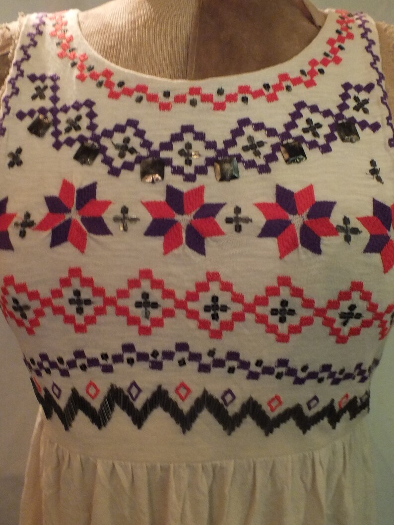May include: A white sleeveless dress with a geometric pattern embroidered in red, purple, and black thread. The dress has a gathered skirt and a round neckline.