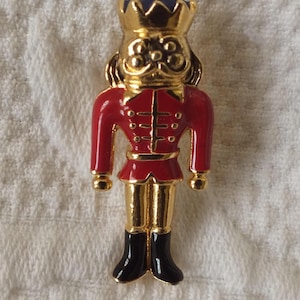 May include: A gold-toned metal brooch in the shape of a nutcracker soldier. The brooch features a red jacket, gold boots, and a blue and gold crown. A small pearl is at the top of the brooch.