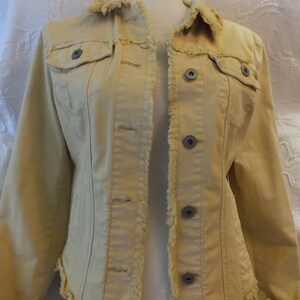 Puede incluir: Chaqueta vaquera amarillo claro con cuello y bordes deshilachados. La chaqueta tiene dos bolsillos en el pecho, botones y una silueta entallada. La tela parece ser un denim suave y de peso medio.