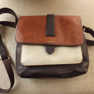 May include: A tri-color Fossil crossbody bag with a brown leather flap, a white front pocket, and a dark brown base. The bag has a long, adjustable strap and a small gold-colored logo.