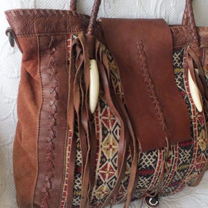 May include: A brown leather tote bag with a suede panel and woven fabric accents. The bag features braided handles, fringe, and decorative bone-like beads. The woven fabric has a geometric pattern in red, black, and gold.