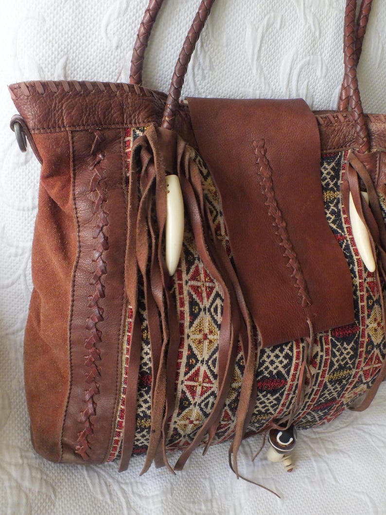 May include: A brown leather tote bag with a suede panel and woven fabric accents. The bag features braided handles, fringe, and decorative bone-like beads. The woven fabric has a geometric pattern in red, black, and gold.