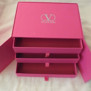 May include: A pink box with three drawers and the Valentino logo on the top. The box is open and the drawers are pulled out.
