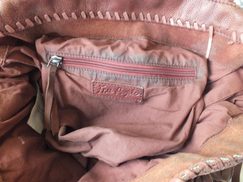 May include: Close-up of a brown leather tote bag interior. The bag features a zippered pocket with a red zipper and a leather tag that reads "Free People". The bag's interior is a dusty rose color.
