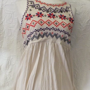 May include: A sleeveless, cream-colored top with a geometric pattern embroidered in red, purple, and black. The top has a gathered skirt and a round neckline.