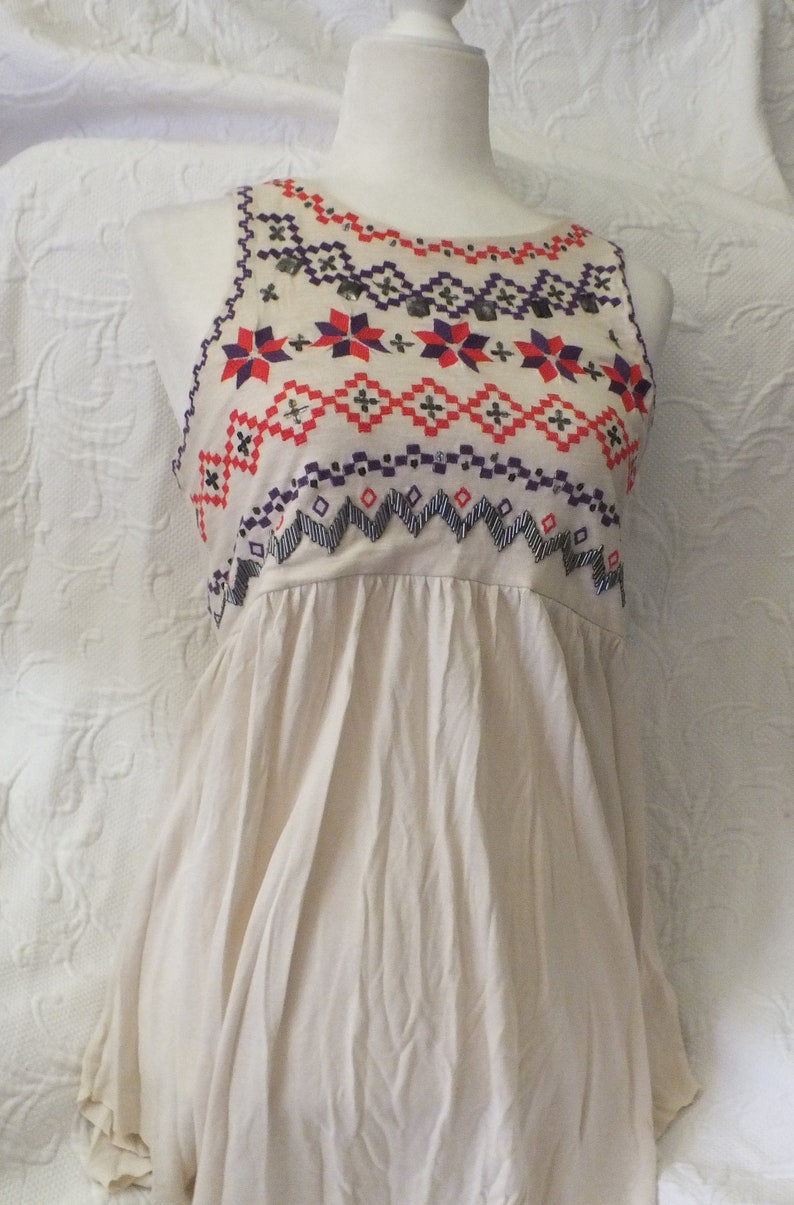 May include: A sleeveless, cream-colored top with a geometric pattern embroidered in red, purple, and black. The top has a gathered skirt and a round neckline.