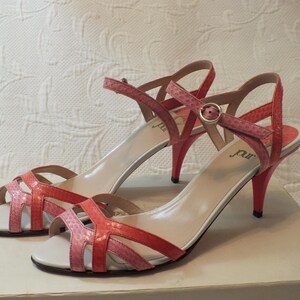 May include: A pair of pink and coral-colored high-heeled sandals. The sandals have a strappy design with a buckle closure and a stiletto heel. The heels are approximately 3 inches high. The sandals are displayed on a white surface.