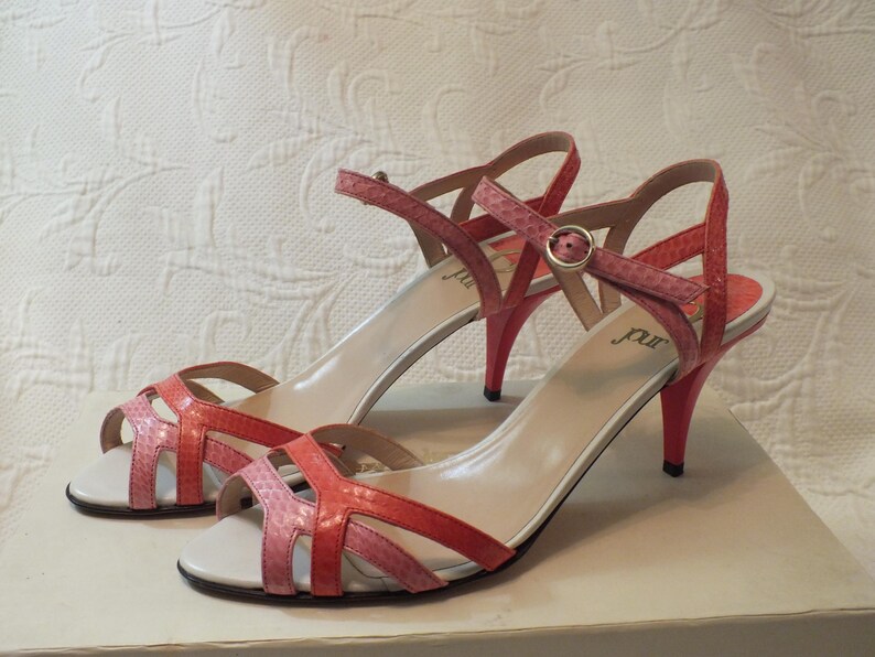 May include: A pair of pink and coral-colored high-heeled sandals. The sandals have a strappy design with a buckle closure and a stiletto heel. The heels are approximately 3 inches high. The sandals are displayed on a white surface.