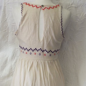 May include: A white sleeveless dress with a geometric pattern embroidered around the neckline and waist. The dress has a keyhole back.