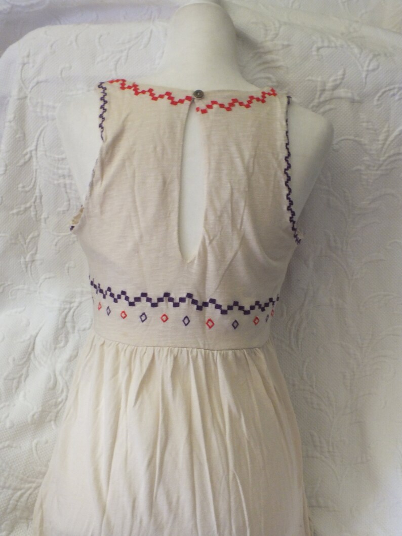 May include: A white sleeveless dress with a geometric pattern embroidered around the neckline and waist. The dress has a keyhole back.
