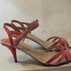 May include: A pair of coral and pink heeled sandals. The sandals have a strappy design with an ankle strap and a small, pointed heel. The shoes are displayed on a white surface.
