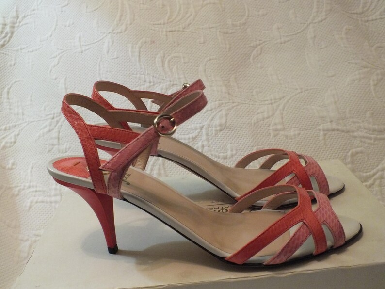 May include: A pair of coral and pink heeled sandals. The sandals have a strappy design with an ankle strap and a small, pointed heel. The shoes are displayed on a white surface.