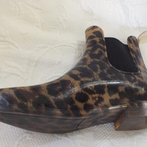 May include: A brown and black leopard print Chelsea boot. The boot has a low heel and a black elastic panel on the side. The pull tab is a clear plastic. The boot is made of a shiny material.
