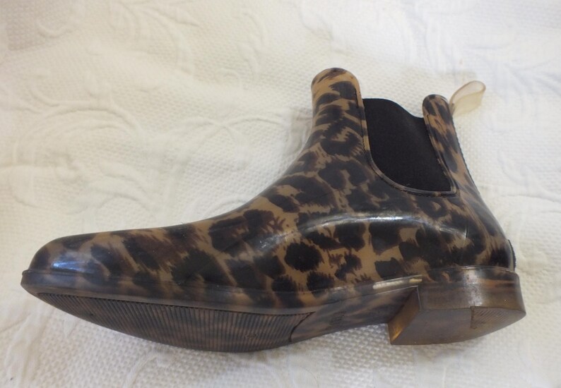 May include: A brown and black leopard print Chelsea boot. The boot has a low heel and a black elastic panel on the side. The pull tab is a clear plastic. The boot is made of a shiny material.