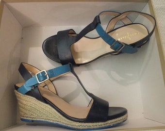 Puede incluir: Dos pares de sandalias de cuña negras con correas y hebillas azules. Las sandalias tienen una plataforma de yute tejido y una parte superior de cuero. El nombre de la marca "Corthay" es visible en la plantilla de una sandalia.