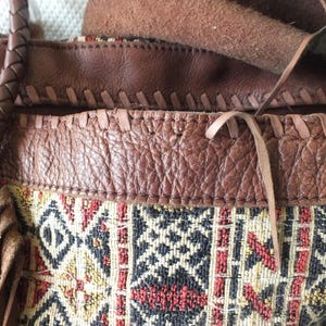 May include: Close-up of a brown leather handbag with a woven fabric panel. The bag features a braided handle and fringe details. The fabric panel has a geometric pattern in red, black, and yellow.