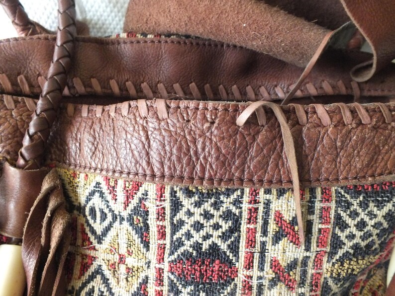 May include: Close-up of a brown leather handbag with a woven fabric panel. The bag features a braided handle and fringe details. The fabric panel has a geometric pattern in red, black, and yellow.