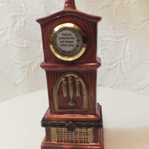 May include: A porcelain clock shaped like a grandfather clock with a gold-colored face and a brown wooden finish. The clock face has a gold-colored rim and the text "Remove protective film and stopper before using".