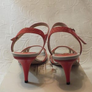 May include: A pair of coral-colored heeled sandals with thin straps. The heels are approximately 3 inches tall. The sandals are displayed on a Coach box, with the brand's logo visible.