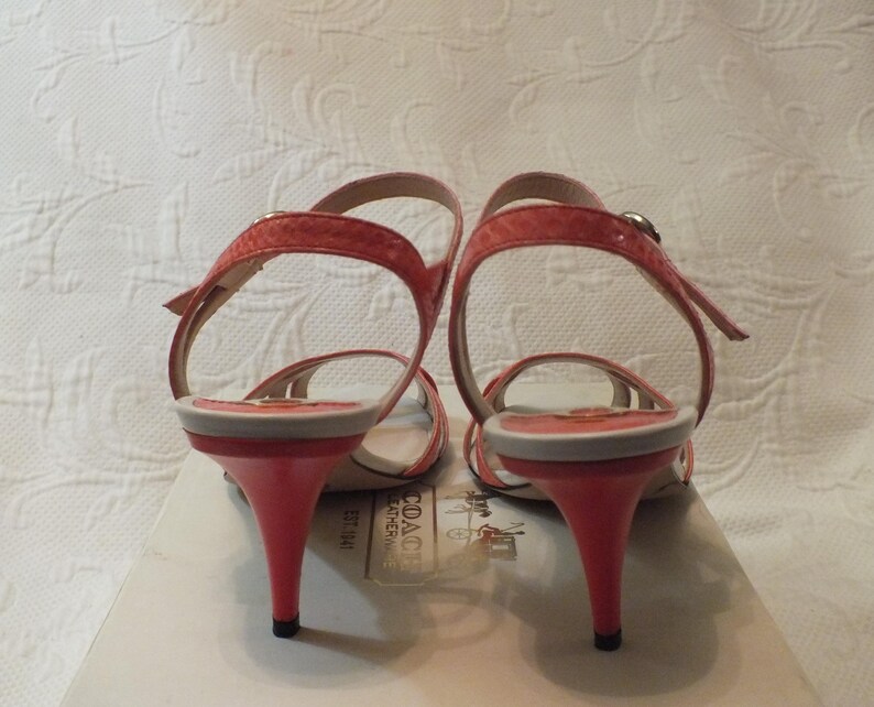 May include: A pair of coral-colored heeled sandals with thin straps. The heels are approximately 3 inches tall. The sandals are displayed on a Coach box, with the brand's logo visible.