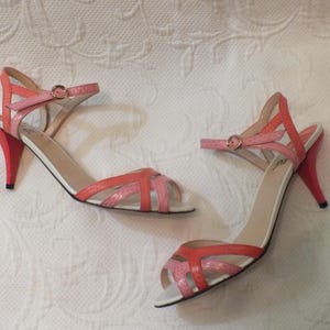 May include: A pair of open-toe sandals with a strappy design in shades of pink and coral. The heels are a vibrant red, and the ankle straps have small gold buckles. The shoes have white soles.