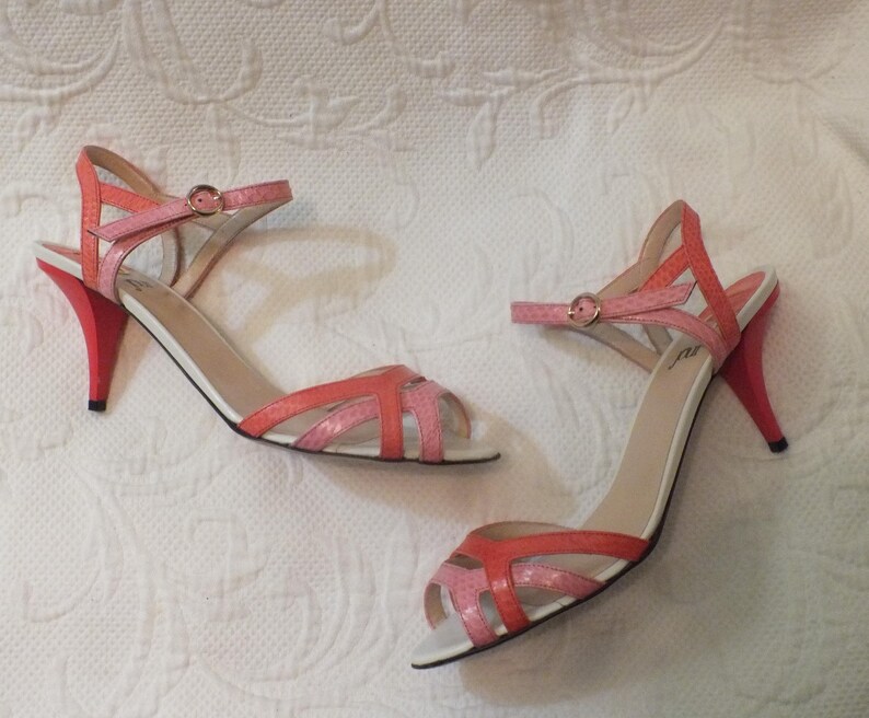 May include: A pair of open-toe sandals with a strappy design in shades of pink and coral. The heels are a vibrant red, and the ankle straps have small gold buckles. The shoes have white soles.