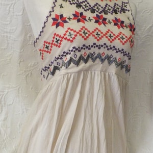 May include: A sleeveless, cream-colored top with a geometric pattern embroidered in red, purple, and black. The top has a gathered neckline and a loose, flowing fit.